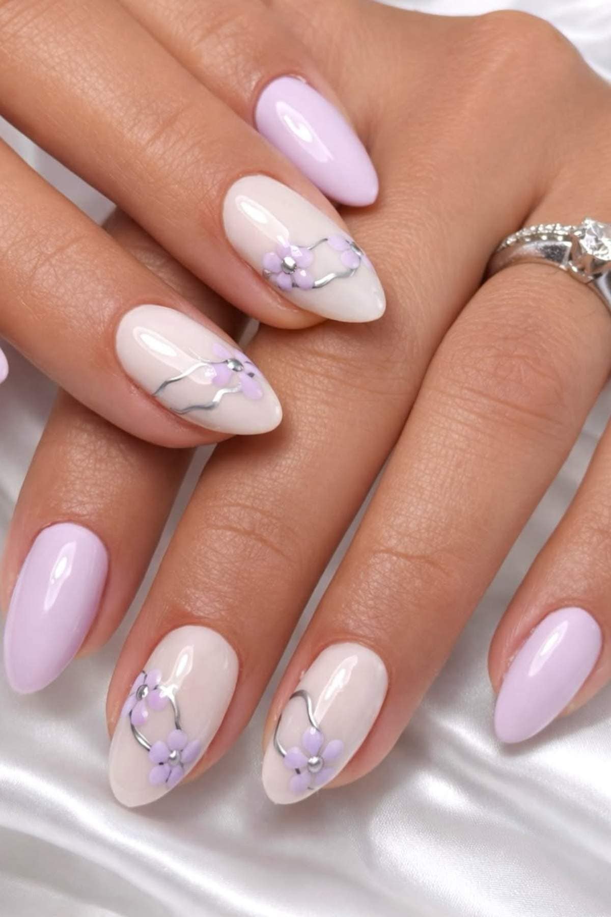 Hands with long almond-shaped nails painted in pale pink and white, featuring small purple floral designs and a silver ring on one finger.