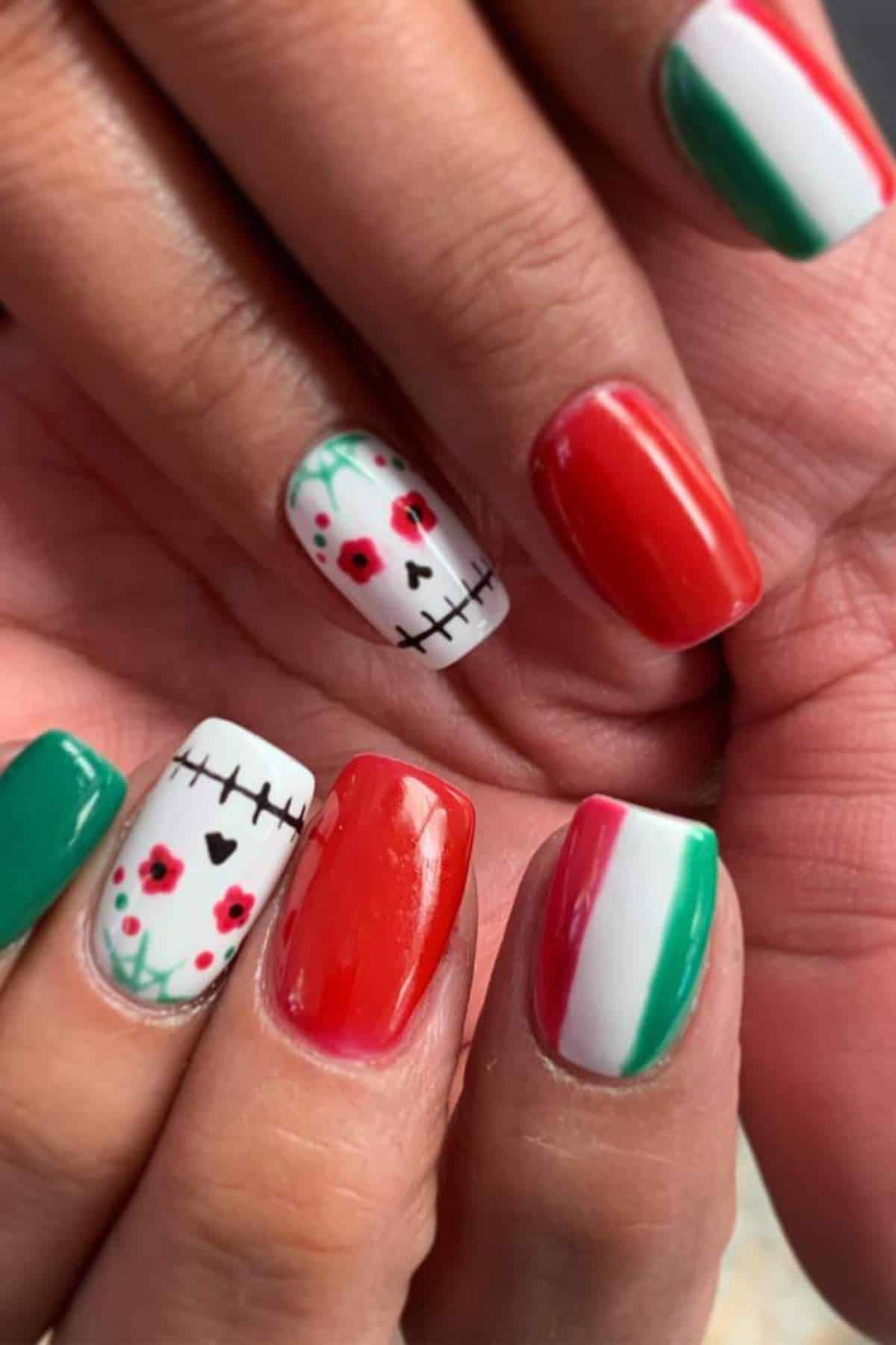 Fingernails painted with red, white, and green colors, some with skull and flower designs resembling Día de los Muertos and Mexican flag themes.
