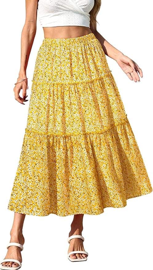 A woman wearing a white crop top, a yellow floral tiered maxi skirt, and white sandals, standing with one arm slightly raised.