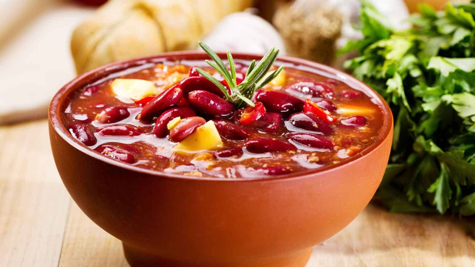A bowl of kidney bean stew garnished with a sprig of rosemary, with herbs and bread in the background.