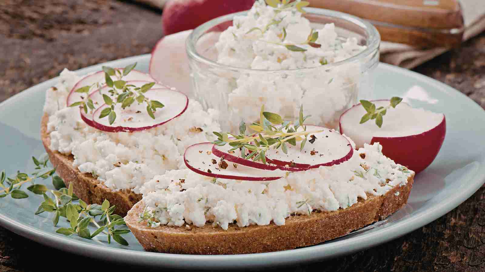 Slices of bread topped with cottage cheese, radish slices, black pepper, and fresh herbs are served on a plate with a small bowl of cottage cheese in the background.