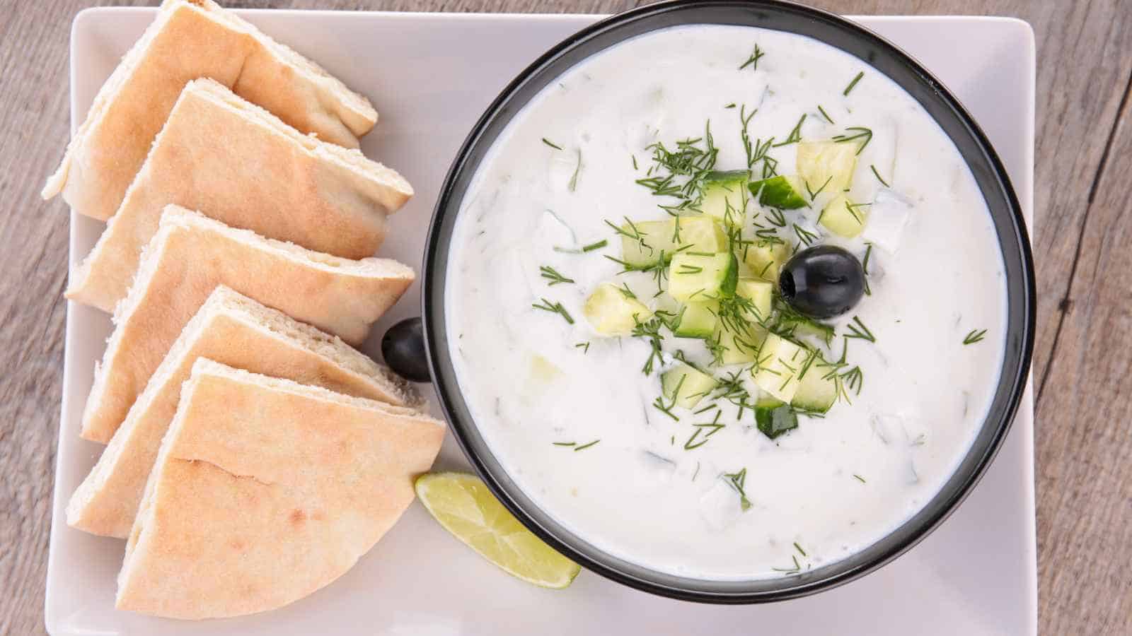A bowl of tzatziki garnished with chopped cucumber, dill, and a black olive, served with pita bread slices and a lime wedge on a white plate.