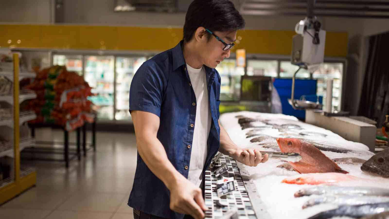 A man wearing glasses examines a fish at a seafood counter in a grocery store, surrounded by various types of fish displayed on ice.