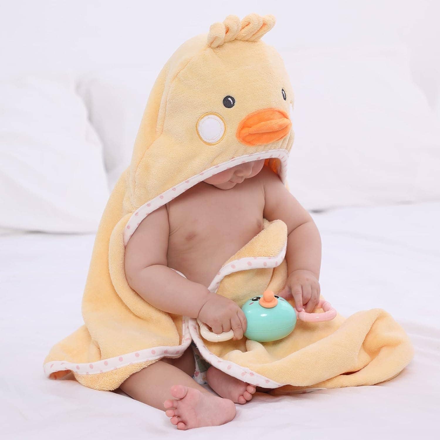 A baby sits on a bed wearing a yellow towel with a duck hood, holding a green and pink toy.