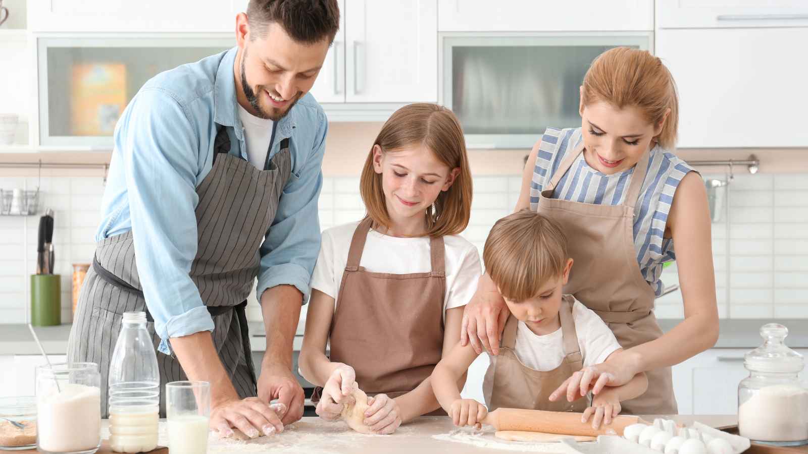 A man, woman, and two children wearing aprons are baking together in a bright kitchen, rolling dough on a floured countertop with ingredients and utensils visible.