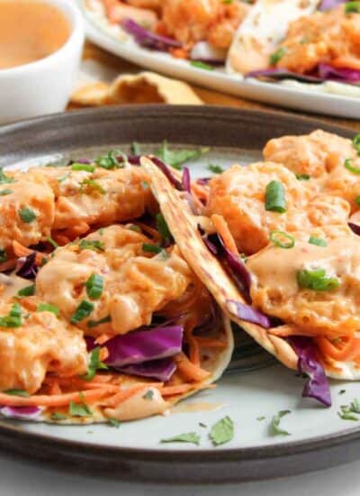A plate of shrimp tacos with shredded purple cabbage, carrots, chopped green onions, and creamy orange sauce, with more tacos and sauce in the background.