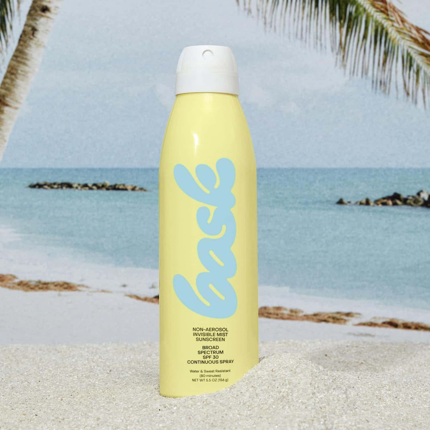 A yellow bottle of Bask non-aerosol sunscreen SPF 30 stands upright in the sand on a beach with the ocean and palm trees in the background.