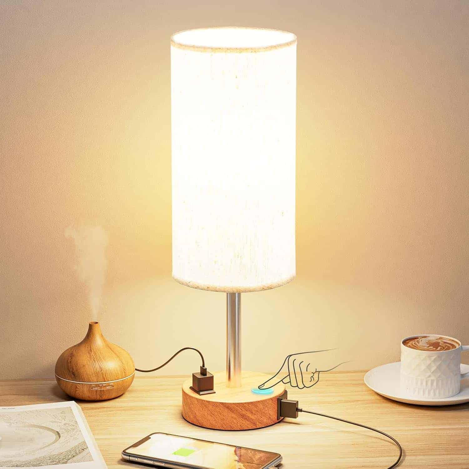 A table lamp with a cylindrical shade features a touch control on its wooden base, two USB ports charging devices, and sits beside a cup of coffee, an aroma diffuser, and a magazine.
