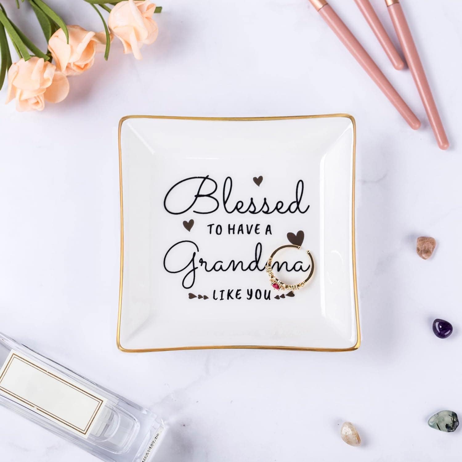 Square ceramic dish with the message "Blessed to have a grandma like you," surrounded by small hearts. Dish is on a white surface with flowers, pens, and decorative stones nearby.