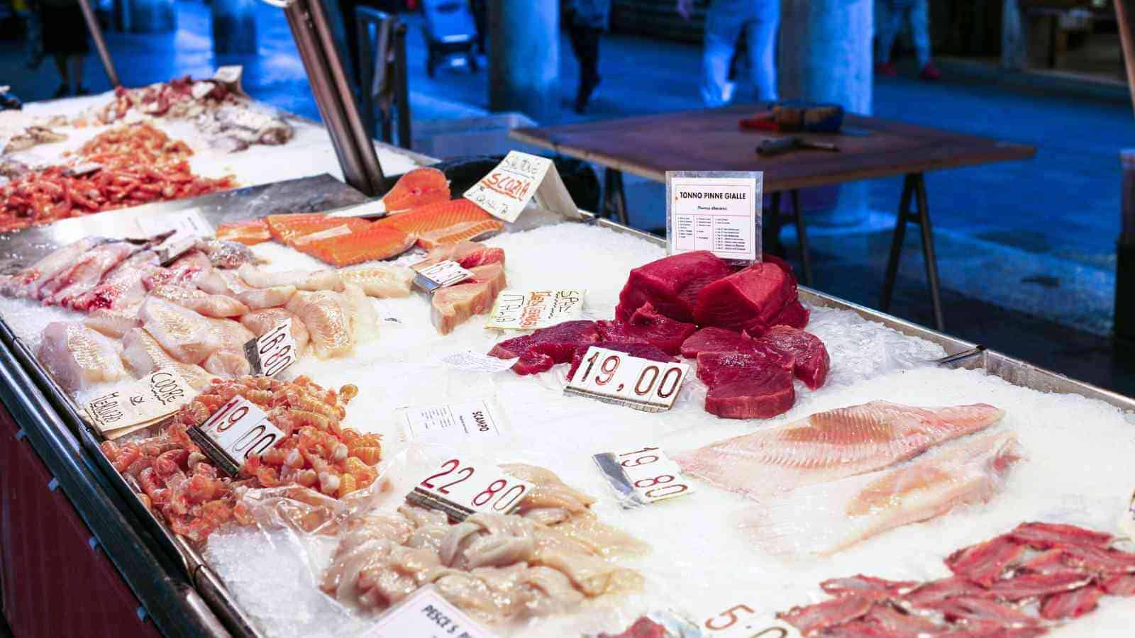Various types of fresh fish and seafood are displayed on ice at a market stall, each with visible price tags.