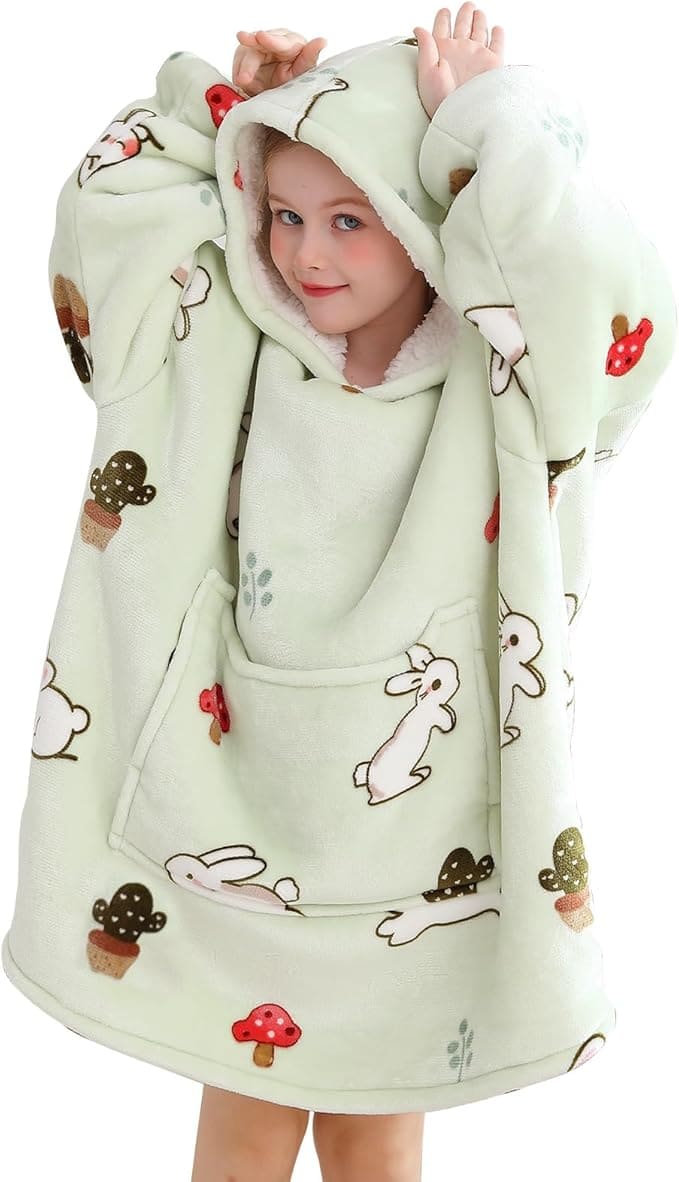Child wearing an oversized, hooded blanket with bunny, mushroom, and cactus patterns, holding up the hood with both hands and smiling.