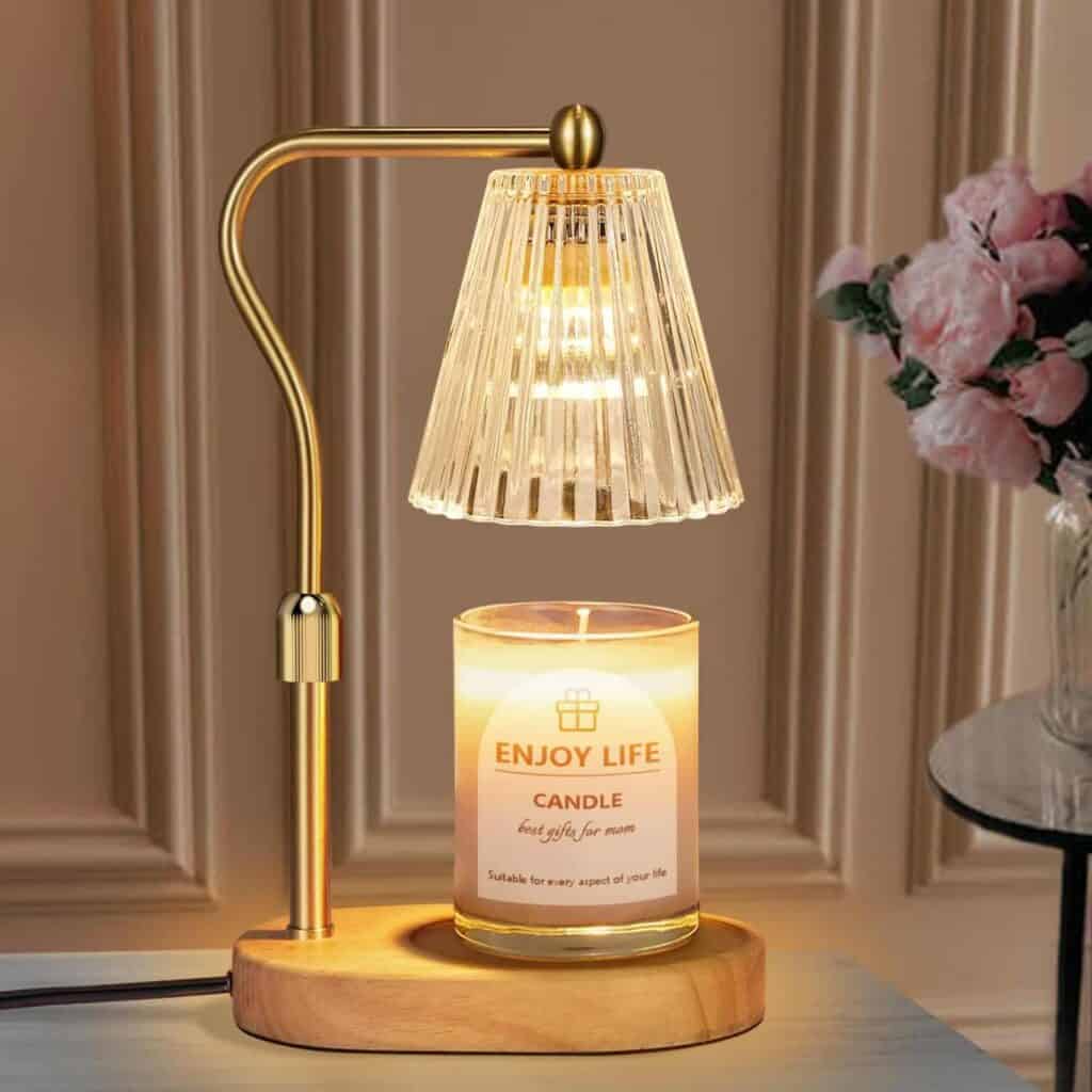 A gold and wood table lamp with a glass shade is positioned above a lit "Enjoy Life" candle, next to a vase of pink flowers on a table.