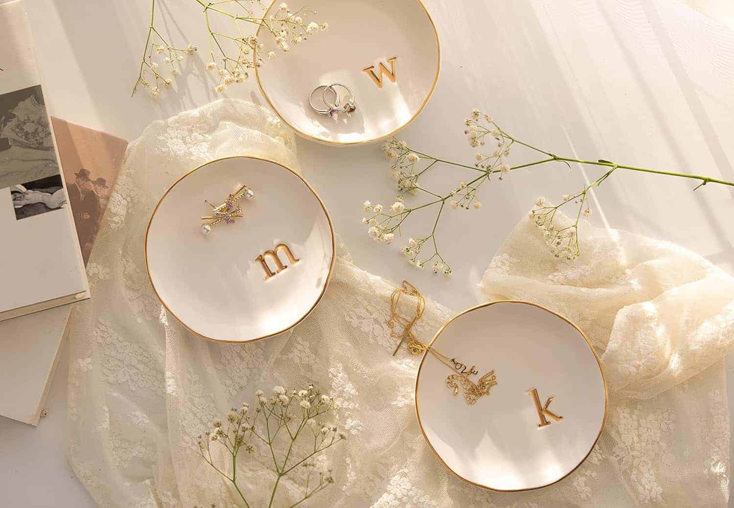 Three white dishes with gold rims hold jewelry and gold letter charms "w," "m," and "k," arranged on a lace fabric with sprigs of baby's breath flowers nearby.