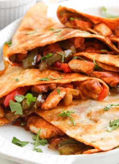 A plate of chicken quesadillas filled with sautéed peppers and onions, garnished with fresh cilantro, sits on a white surface.