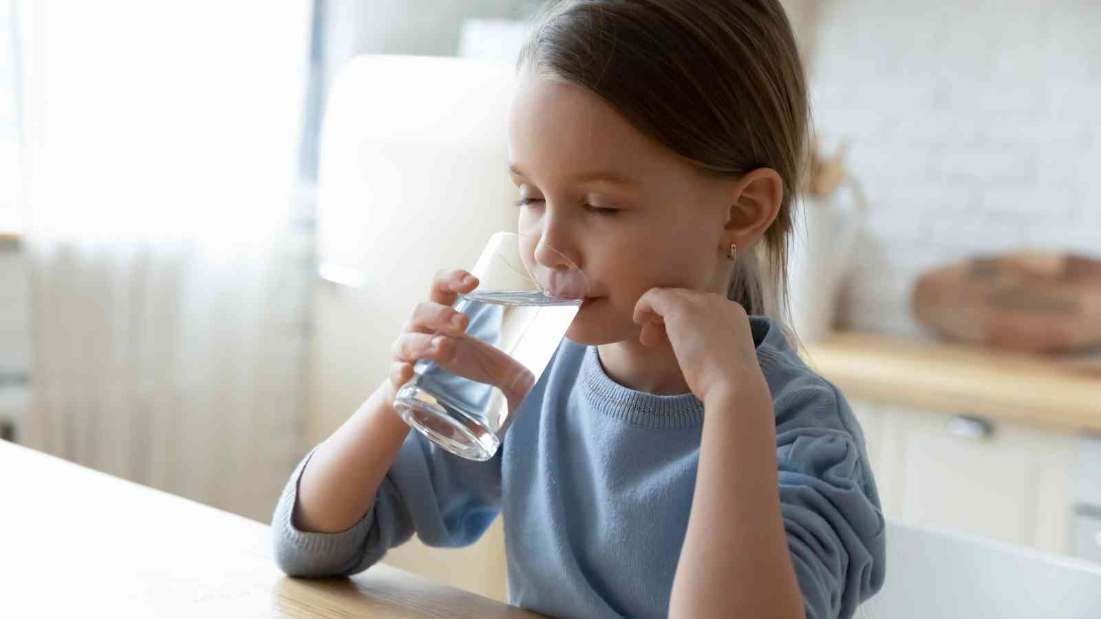 A young girl with brown hair drinks a glass of water while sitting at a kitchen table.