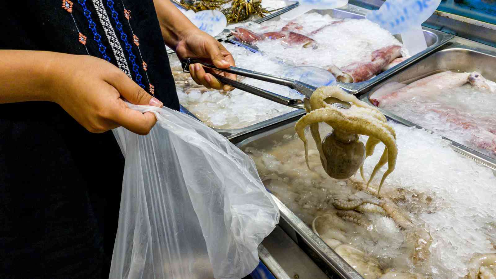 A person uses tongs to pick up a raw octopus from an ice-filled seafood display and places it in a plastic bag.