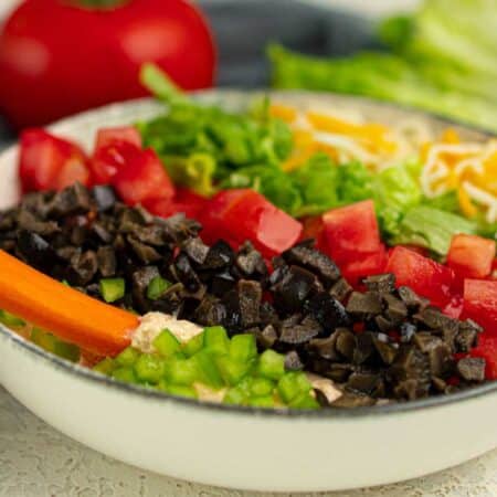 A bowl of layered cottage cheese dip topped with chopped tomatoes, shredded cheese, lettuce, olives, green onions, and a carrot stick.