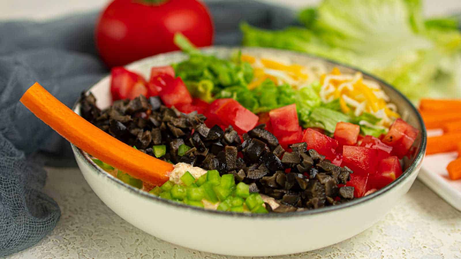 A bowl of layered cottage cheese dip topped with chopped tomatoes, shredded cheese, lettuce, olives, green onions, and a carrot stick.