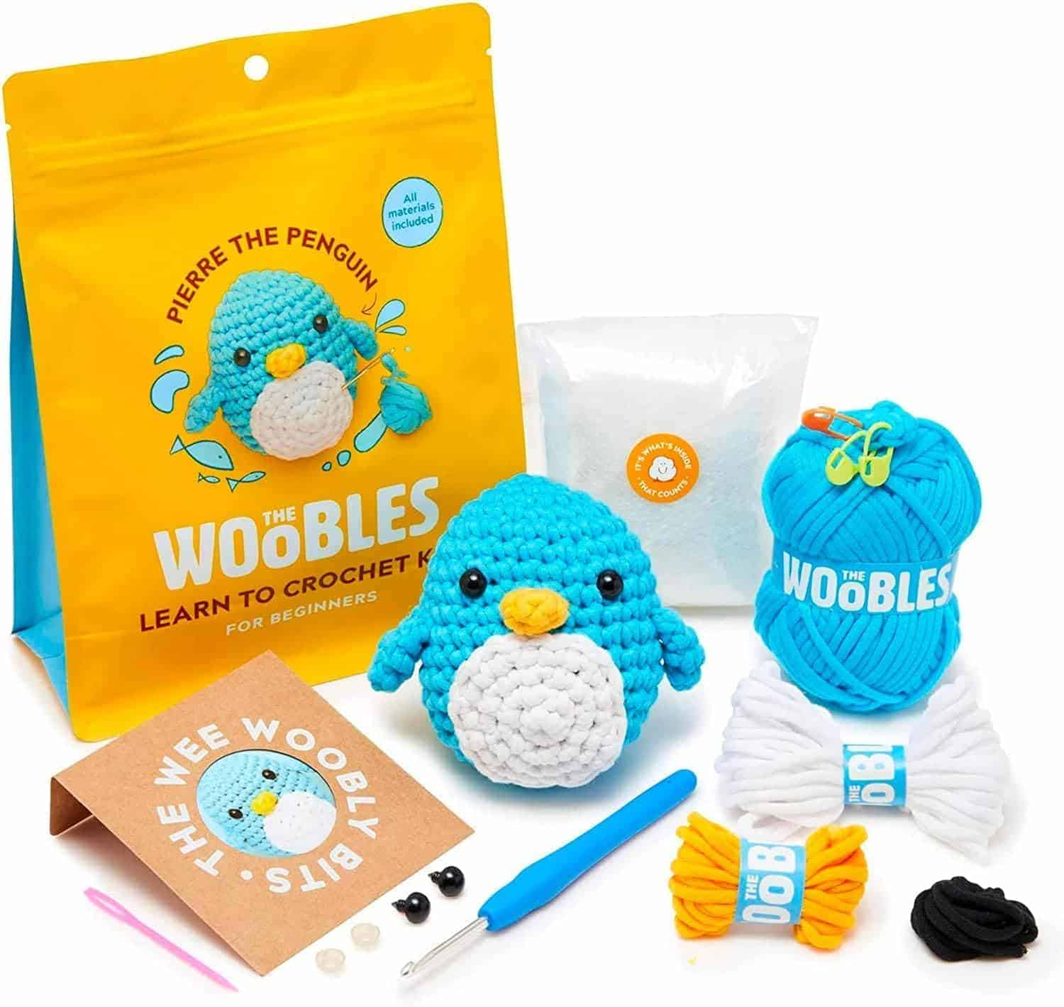 Beginner crochet kit featuring a blue penguin, yarn, crochet hook, needle, eyes, stuffing, instruction booklet, and a yellow bag labeled "The Woobles: Learn to Crochet Kit.