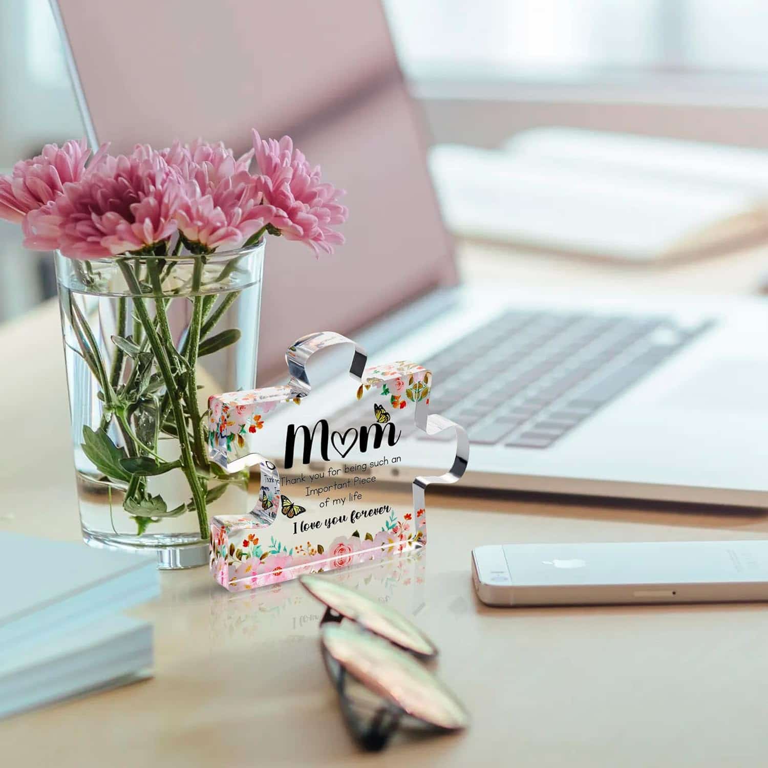 A glass vase with pink flowers, a laptop, a smartphone, and a decorative puzzle piece with a "Mom" message sit on a white desk.