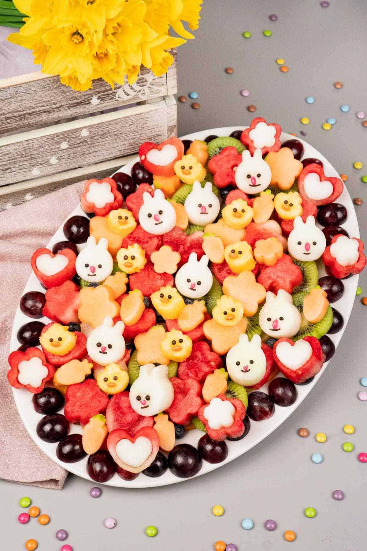 An oval Easter platter with fruit pieces cut into shapes of bunnies, chicks, hearts, and flowers, surrounded by grapes, with yellow flowers and colorful candy on the table.