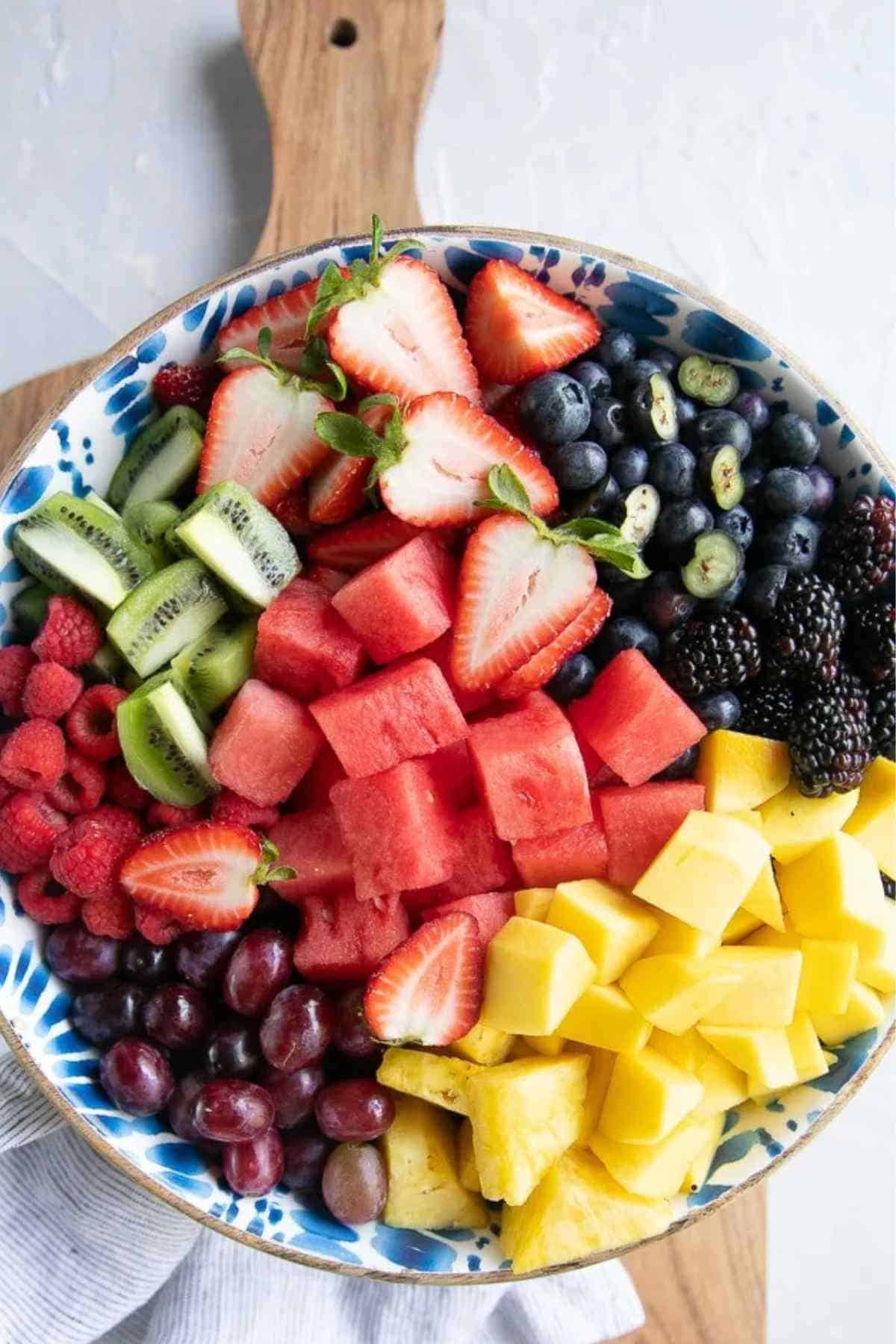 A bowl filled with sliced strawberries, blueberries, blackberries, kiwi, raspberries, watermelon, grapes, mango, and pineapple on a wooden board.
