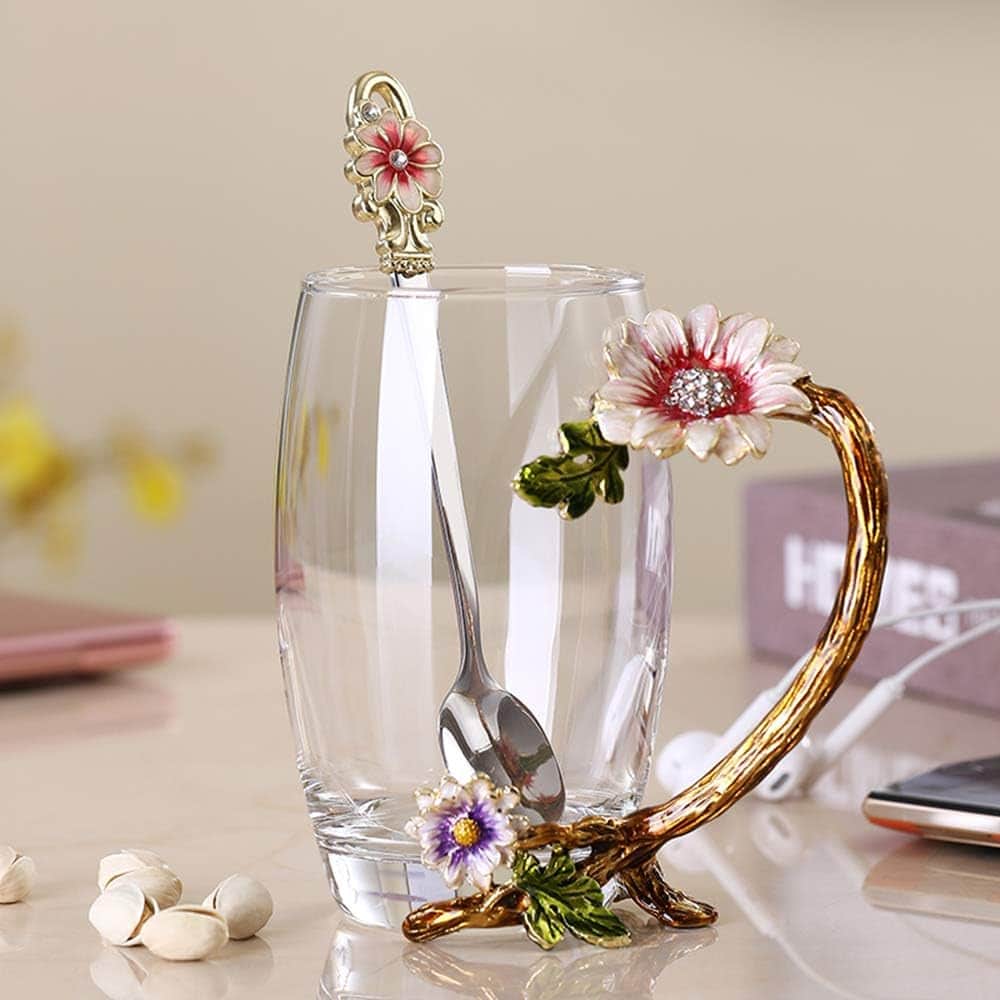 A clear glass with a decorative flower handle and a matching floral spoon is placed on a table next to pistachios, a book, and white earphones.
