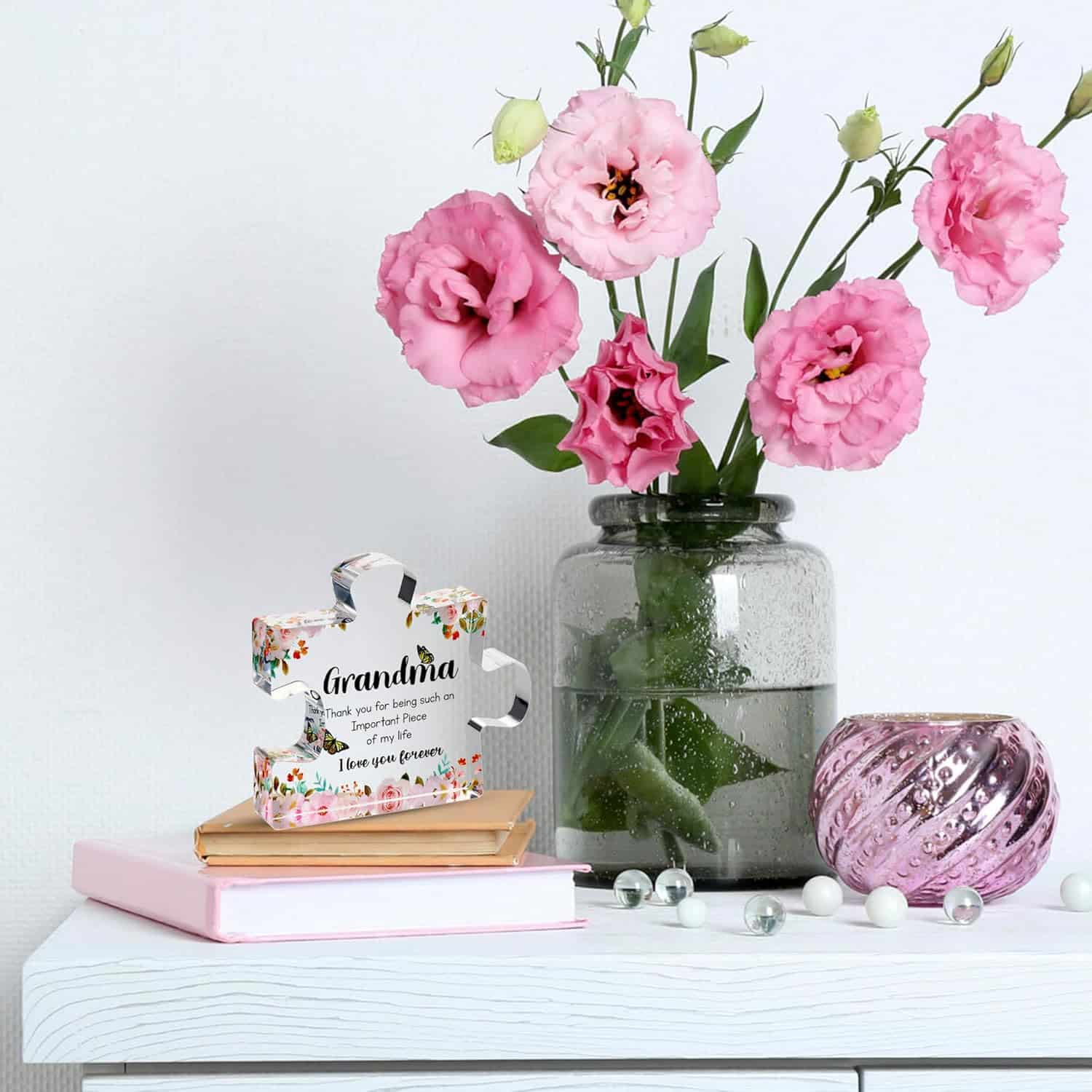 A glass vase with pink flowers, a glass ornament, marbles, and a decorative puzzle piece with a message for Grandma are arranged on a white table with stacked books.