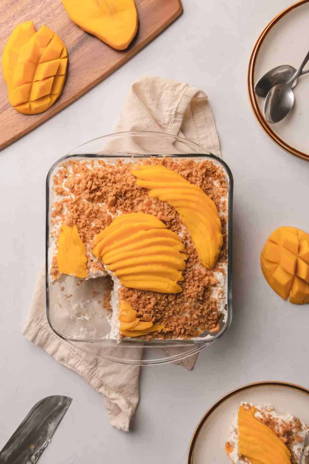 A glass dish of mango float dessert topped with sliced mangoes and crumbs, with a serving removed. Sliced mangoes and spoons are on the side.