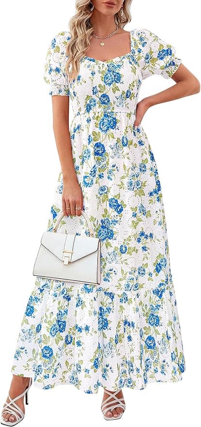 A woman wearing a white maxi dress with blue and green floral print holds a white handbag and stands in white heeled sandals.