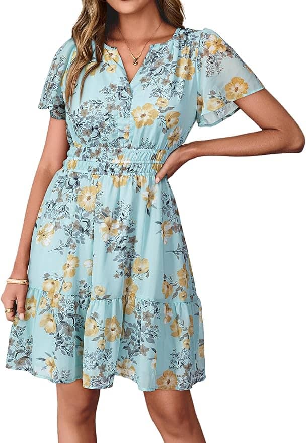 A woman wearing a light blue, short-sleeve, floral print dress with yellow and gray flowers, standing with one hand on her hip.