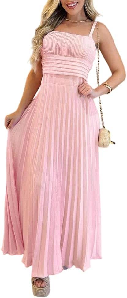 A woman wearing a sleeveless, light pink pleated maxi dress, holding a small beige purse and standing in wedge sandals.