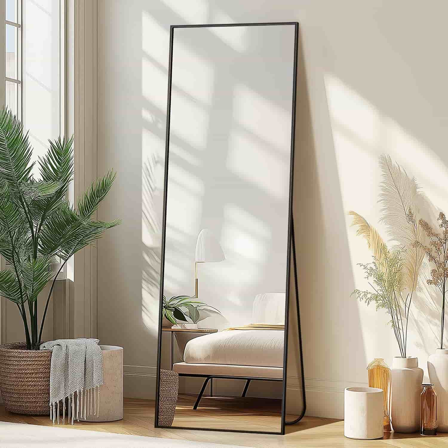 A tall, rectangular mirror with a thin black frame leans against a wall in a bright, modern bedroom with potted plants and neutral decor.