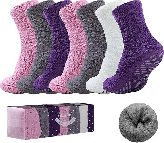 A set of six pairs of fuzzy, non-slip socks in purple, pink, gray, and white colors, displayed standing upright with packaging and a rolled-up sock shown in front.