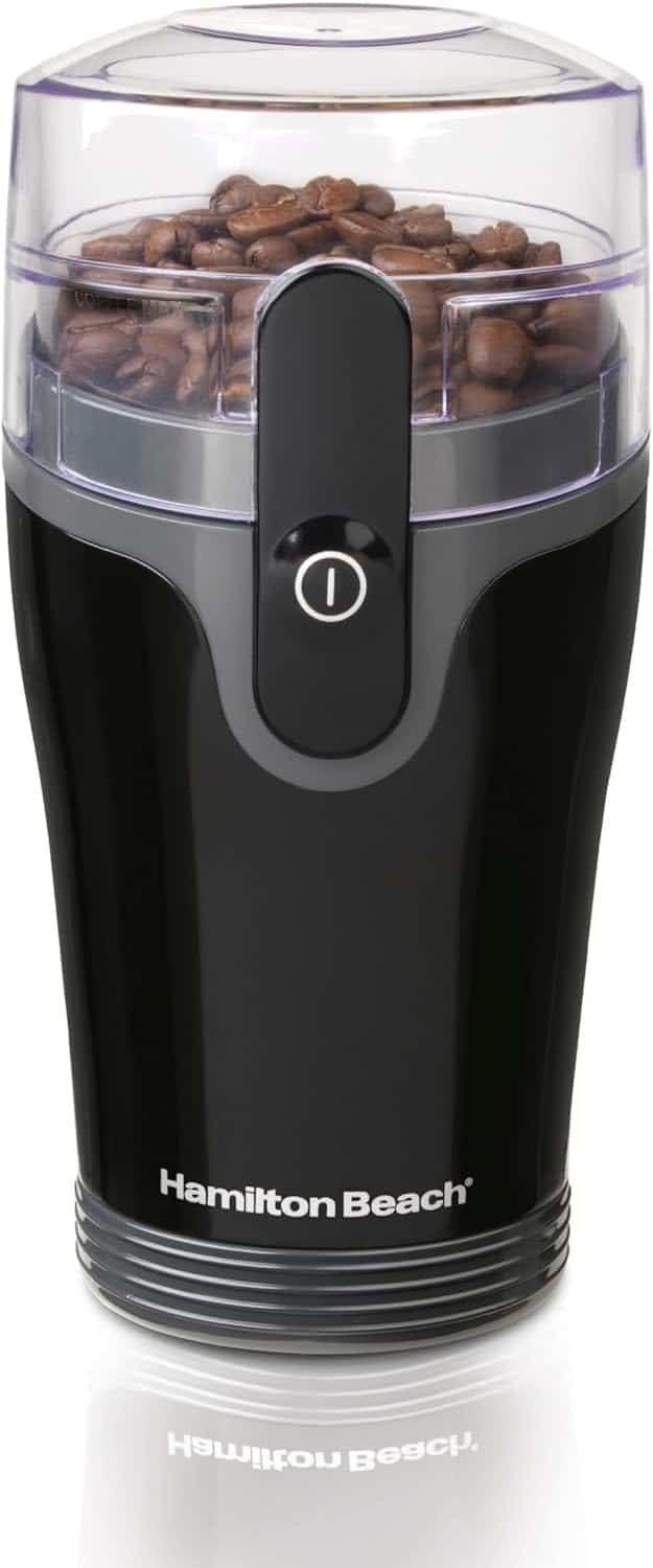 A black Hamilton Beach electric coffee grinder with coffee beans inside, shown with a transparent lid and a central power button.