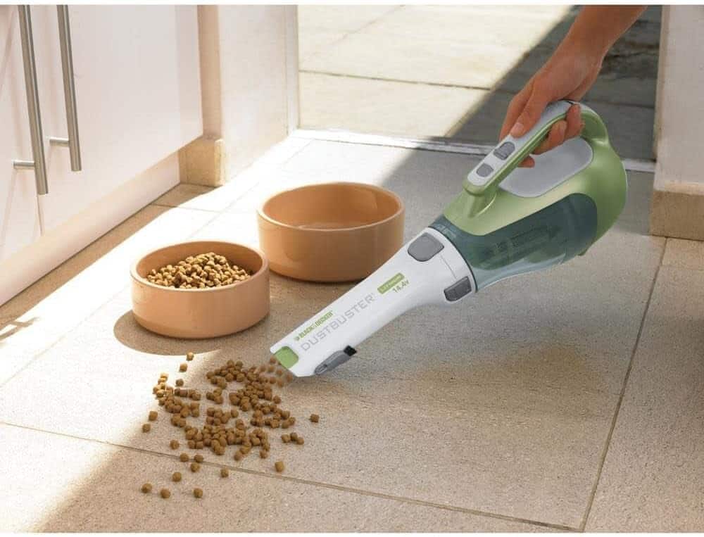 A person uses a handheld vacuum to clean up spilled pet food next to two bowls on a tiled floor.