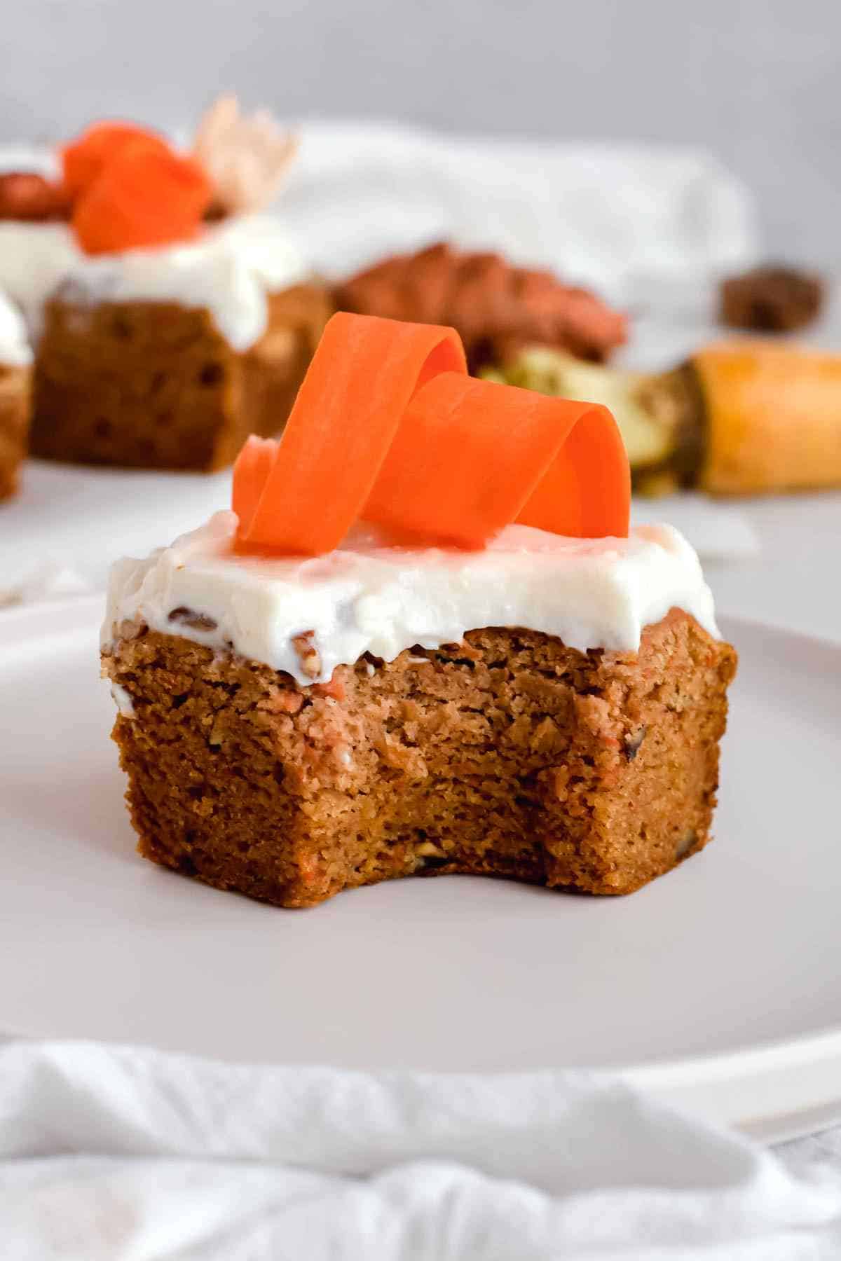 A square piece of carrot cake with white frosting and carrot ribbons on top, served on a white plate with a bite taken out.