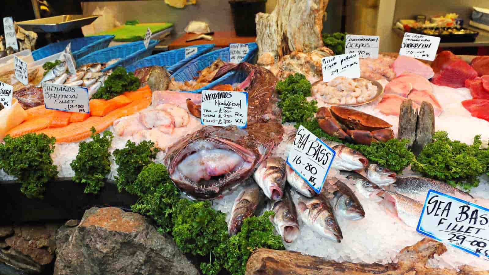 A seafood market display featuring various fish, shellfish, and fillets on ice, with handwritten price signs and garnished with parsley.