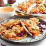 A plate of Bang Bang shrimp tacos with shredded purple cabbage, carrots, chopped green onions, and creamy orange sauce, with more tacos and sauce in the background.