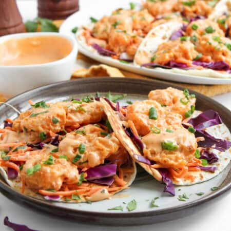 A plate of Bang Bang shrimp tacos with shredded purple cabbage, carrots, chopped green onions, and creamy orange sauce, with more tacos and sauce in the background.