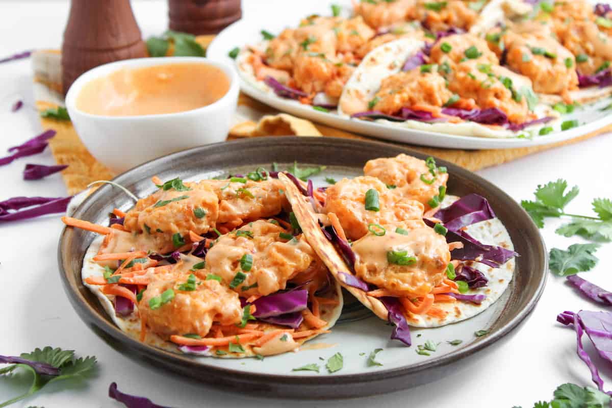 A plate of shrimp tacos with shredded purple cabbage, carrots, chopped green onions, and creamy orange sauce, with more tacos and sauce in the background.