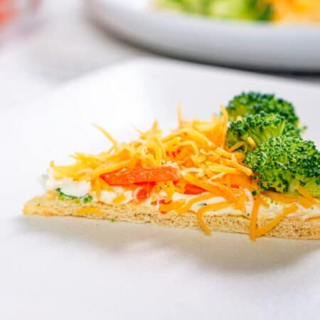 A slice of veggie pizza topped with shredded cheddar cheese, broccoli florets, and red bell pepper on a square white plate.