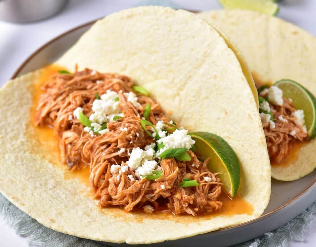 Two soft tortillas filled with shredded chicken in sauce, topped with crumbled cheese, chopped green onions, and lime wedges on a plate.