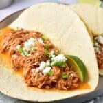 Two soft tortillas filled with shredded chicken in sauce, topped with crumbled cheese, chopped green onions, and lime wedges on a plate.