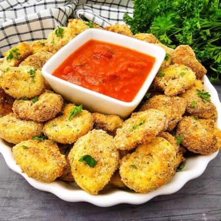 A plate of breaded parmesan tortellini bites with parsley, served with a bowl of red marinara sauce, with a checkered towel and fresh parsley in the background.