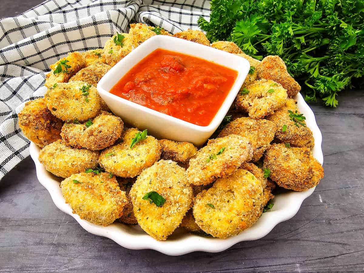 A plate of breaded, fried zucchini slices garnished with parsley, served with a bowl of red marinara sauce, with a checkered towel and fresh parsley in the background.