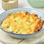 A baked cheesy spinach artichoke dip in a blue bowl, placed on a round marble board with vegetables and kitchen items in the background.