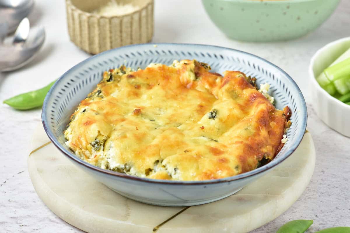 A baked cheesy spinach dip in a blue bowl, placed on a round marble board with vegetables and kitchen items in the background.