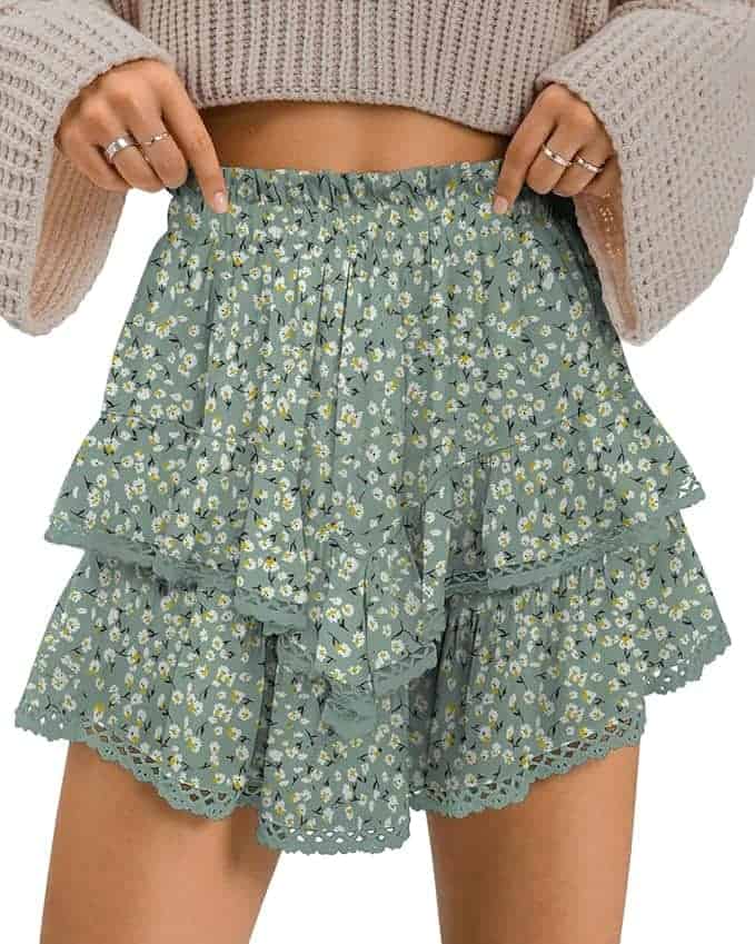 A person wearing a beige knit sweater and a green ruffled mini skirt with a small white floral pattern and lace trim, pointing at the waistband.