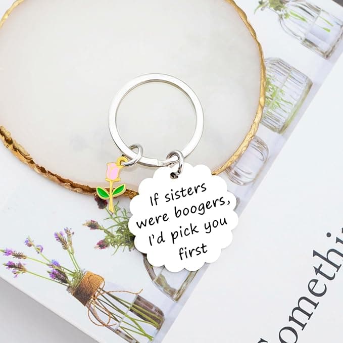 A keychain with a rose charm and a scalloped tag that reads, "If sisters were boogers, I’d pick you first," placed on a book with a floral cover.